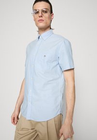 Camisa azul clara de manga curta com botões, com bolso no peito e bainha curvada, feita de tecido texturizado. Botões em aço inoxidável acrescentam detalhe.