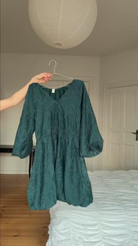 Roka tur zaļu kleitu ar ziedu rakstu un trim ceturtdaļas piedurknēm uz pakaramā guļamistabā ar baltu gultu un koka grīdu.