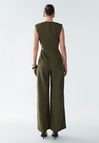 Salopette vert olive avec un corsage ajusté, des jambes larges et des manches courtes. Présente une fermeture éclair au dos et une texture lisse et mate.
