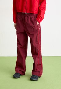 Maroon lagane hlače s elastičnom trakom u struku, bočnim džepovima i kontrastnim crvenim šavovima. Nošene s crnim cipelama na zelenoj površini.