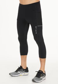 Schwarze sportliche Leggings mit einer figurnahen Passform, die ein strukturiertes Panel an der Seite und ein reflektierendes Logo aufweisen. Mit einer Länge bis zur Mitte des Waden.