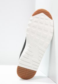 Svart sneaker med texturerad ovandel, vit gummisula med rutmönster och en brun gummidetalj på hälen.