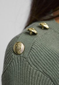 Groene geribbelde trui met vier gouden knopen op de schouder, voorzien van een textuurstof en een aansluitend design.