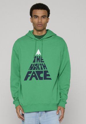 Muž v zelené mikině s kapucí s trojúhelníkovým logem "The North Face" v tmavě modré a bílé barvě, stojící před jednoduchým světlým pozadím.