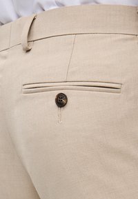 Pantaloni beige su misura con fronte piatto, tasca posteriore con bottone scuro e un sottile tessuto testurizzato.