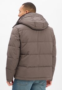 Mann in einer braunen gepolsterten Jacke mit Kapuze, von hinten betrachtet, kombiniert mit blauen Jeans vor einem schlichten Hintergrund.