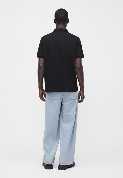 Homme portant un polo noir à manches courtes, un jean bleu clair à jambes larges et des chaussures noires, debout de dos sur un fond uni.