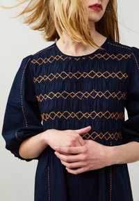 Navyblaues Kleid mit Puffärmeln, das orangene gestickte Diamantmuster über dem Oberteil und einen fein strukturierten Stoff aufweist.