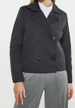 Frau trägt eine schwarze, doppelreihige Jacke über einem weißen Hemd mit Kragen und grauen, maßgeschneiderten Hosen vor einem schlichten Hintergrund.