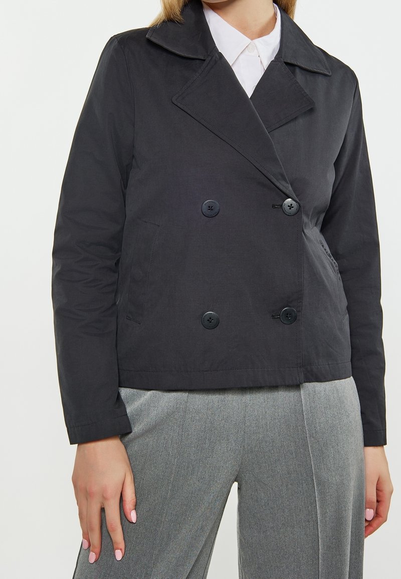 Femme portant une veste noire croisée sur une chemise blanche à col et un pantalon gris sur mesure sur un fond uni.