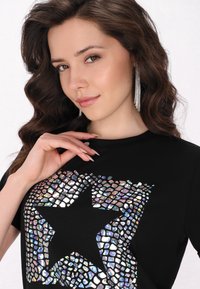 Černé tričko s velkým, reflexním designem hvězdy vyrobené z fragmentovaných, iridescentních materiálů. Hladký textilní povrch, krátké rukávy.