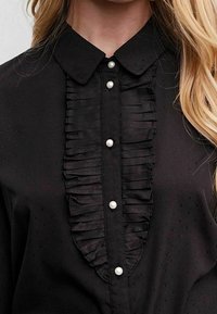 Blusa nera con pannello frontale arricciato e tre bottoni in perla, colletto classico e una sottile texture nel tessuto.