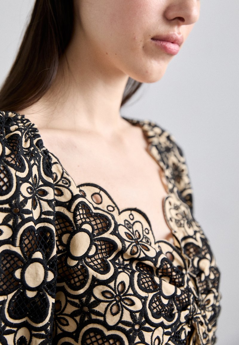 Black floral lace overlay on a beige fabric, featuring a scalloped neckline and puff sleeves, highlighting the intricate floral patterns.