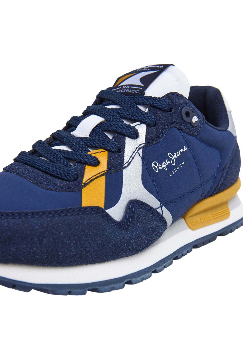 Sneaker blu navy con un mix di materiale tessile e suede, accenti bianchi e gialli, superficie testurizzata e suola bianca. Logo del marchio visibile.