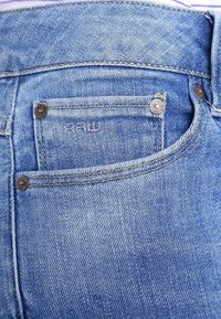 Vaqueros de mezclilla azul con un bolsillo trasero que presenta detalles de costura, con la marca "RAW" y remaches metálicos para mayor durabilidad.