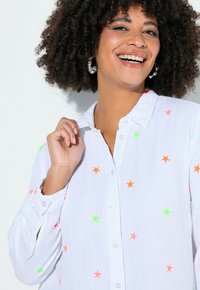 Lächelnde Frau mit lockigem Haar trägt Perlen-Creolen und ein weißes Hemd mit Knöpfen, das mit neongrünen und orangen Sternen verziert ist.