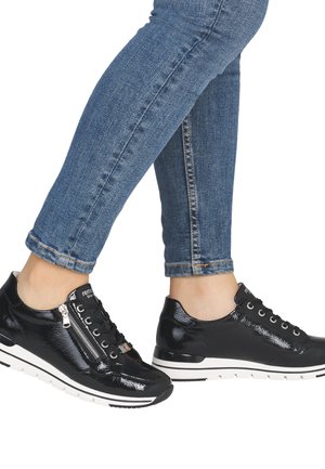Person trägt dunkelblaue Skinny-Jeans und glänzende schwarze Low-Top-Sneaker mit weißen Sohlen und seitlichen Reißverschlüssen.