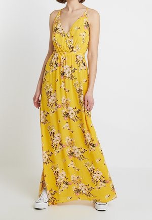 Frau trägt ein gelbes, ärmelloses Maxikleid mit Blumenmuster, Spaghettiträgern und weißen Turnschuhen vor einem einfarbigen Hintergrund.