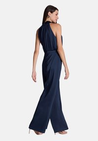 Combinaison en marine avec un col halter, des jambes larges et une taille cintrée. Fabriquée en tissu lisse, elle est dotée d'une fermeture éclair au dos.