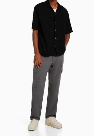 Uomo che indossa una camicia nera a maniche corte con bottoni, pantaloni cargo grigi con tasche e sneakers beige chiaro, in piedi con una mano in tasca.