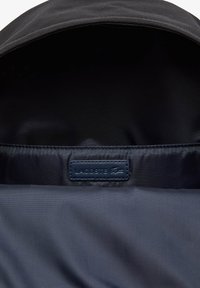 Interior de tela negra con una textura suave, que presenta una etiqueta azul marino con "LACOSTE" y un logo, situada cerca del área del zipper.