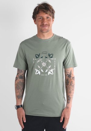 T-shirt vert olive en coton, avec un design graphique central portant le texte "STAY ALIVE" et des motifs de feuilles, manches courtes et col rond.