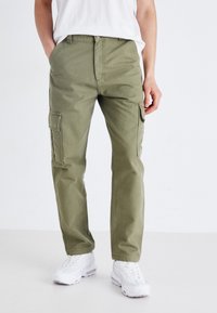 Khaki színű, egyenes szabású cargo nadrág, több oldalsó zsebbel és sima pamut anyagból. Fehér sportcipővel párosítva, kontrasztos élénk színben.