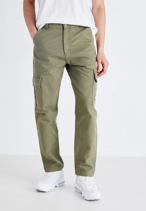 Khaki cargo kalhoty s rovný fit, několika bočními kapsami a hladkým bavlněným materiálem. Doplnené o bílé tenisky, což vytváří kontrastující jasnou barvu.