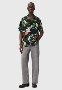 Czarna koszula w kwiaty na krótkie rękawy z czerwonymi, białymi i zielonymi kwiatami, zestawiona z szarymi dżinsami o szerokich nogawkach i czarnym obuwiem.