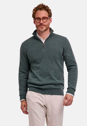 Uomo con capelli ricci e occhiali che indossa un maglione grigio scuro con zip sopra una camicia bianca e pantaloni beige chiaro, in piedi davanti a uno sfondo semplice.