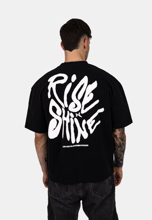 Schwarzes Baumwoll-T-Shirt mit auffälligem weißen Grafiktext "RISE x SHINE" auf dem Rücken. Kurze Ärmel und ein lässiger Schnitt.