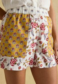 Woman wearing white shorts with elastic waist and mixed floral patterns in red, blue, and yellow, standing with one hand visible.