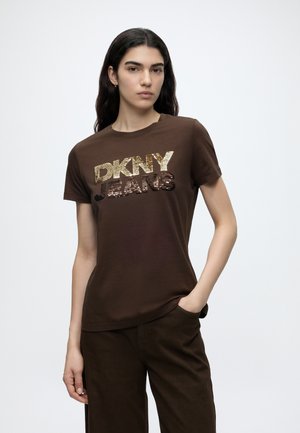 Kobieta z długimi ciemnymi włosami nosi brązową koszulkę z krótkim rękawem z logo "DKNY JEANS" z cekinami w kolorze złotym i ciemnobrązowym oraz pasujące brązowe spodnie.