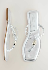 Sandales plates métalliques argentées avec des fines lanières et une petite perle décorative argentée sur fond beige.