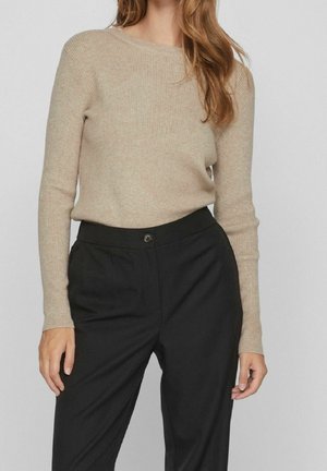 Femme portant un pull beige côtelé à manches longues rentré dans un pantalon noir taille haute avec une fermeture bouton.