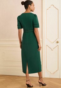 Femme avec les cheveux en chignon portant des boucles d'oreilles dorées et une robe midi vert foncé à manches courtes, avec des talons noirs, se tenant dans une pièce décorative.
