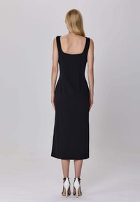 Femme blonde de dos, portant une robe midi noire sans manches avec un décolleté carré et des sandales à talons hauts blanches avec des brides autour de la cheville.
