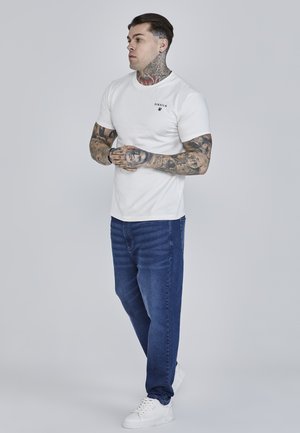 Tricou alb din bumbac cu un mic logo negru, asortat cu blugi din denim albastru și teneși albi. Blugii au detalii subtile de textură și cusături.