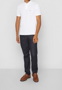 Chemise polo blanche à manches courtes, assortie à un jean bleu foncé et des mocassins marron. La polo présente un petit logo sur la poitrine.