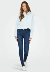 Light blue cropped button-up shirt with long sleeves, paired with dark blue skinny jeans and white sneakers. Smooth fabric and classic design.