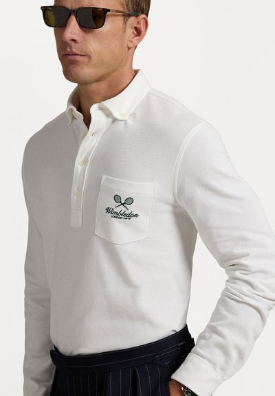Polo alb cu mânecă lungă și un mic buzunar. Are logo-ul brodat "Wimbledon". Poartă-l cu pantaloni în dungi închise și ochelari de soare.