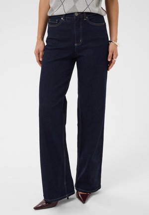 Femme portant un jean large bleu foncé, un pull gris à motif losanges, des chaussures bordeaux pointues et des bijoux en argent aux mains.