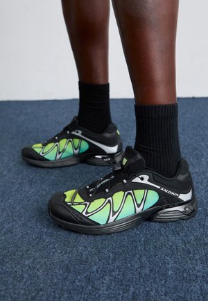 Black and green running shoes with a mesh upper, accented by silver details and a curved sole. Worn with black ankle socks on a blue carpet.