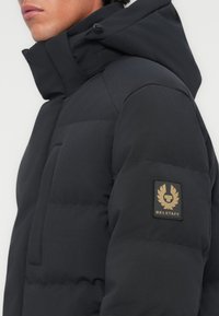 Chaqueta acolchada negra con cuello alto y capucha ajustable. Presenta un bolsillo frontal y un parche con logotipo dorado en la manga izquierda. Textura suave.