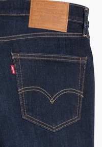 Mörkblå denimjeans med en läderetikett med präglad text, gula sömmar och en distinkt fickdesign.