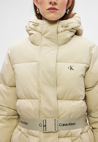 Puffjacka i beige med hög krage, midjebälte och Calvin Klein-logotyp på bröstet. Har sömmade paneler och slät textur.
