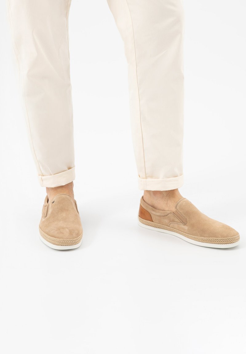 Mann trägt beige Wildleder-Slipper mit weißen Sohlen und beige hochgekrempelte Hosen und steht vor weißem Hintergrund.