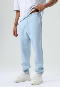 Lichtblauwe joggingbroek van zachte stof, met een elastische tailleband, omgeslagen enkels en zijzakken. Draagt witte sneakers.