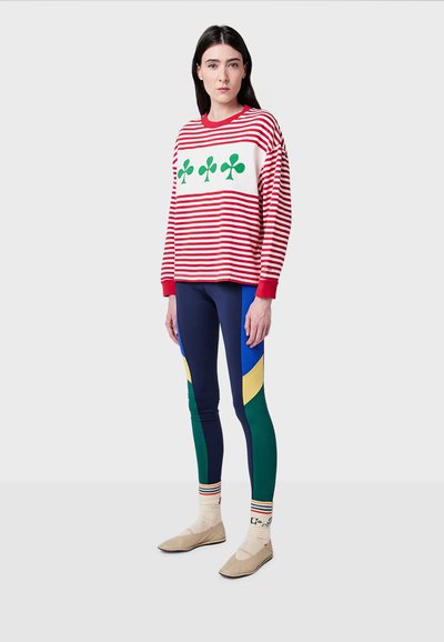 Felpa a righe rosse e bianche con trifogli verdi, abbinata a leggings multicolori con sezioni blu, verdi e gialle. Calzature beige.