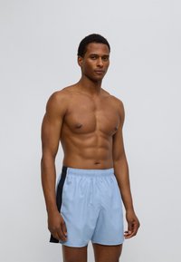 Ανοιχτό μπλε μαγιό βερμούδα με μαύρη πλευρική φάσα, ελαστική μέση και λεία υφή, φορεμένη από μοντέλο χωρίς μπλούζα σε ουδέτερο φόντο.